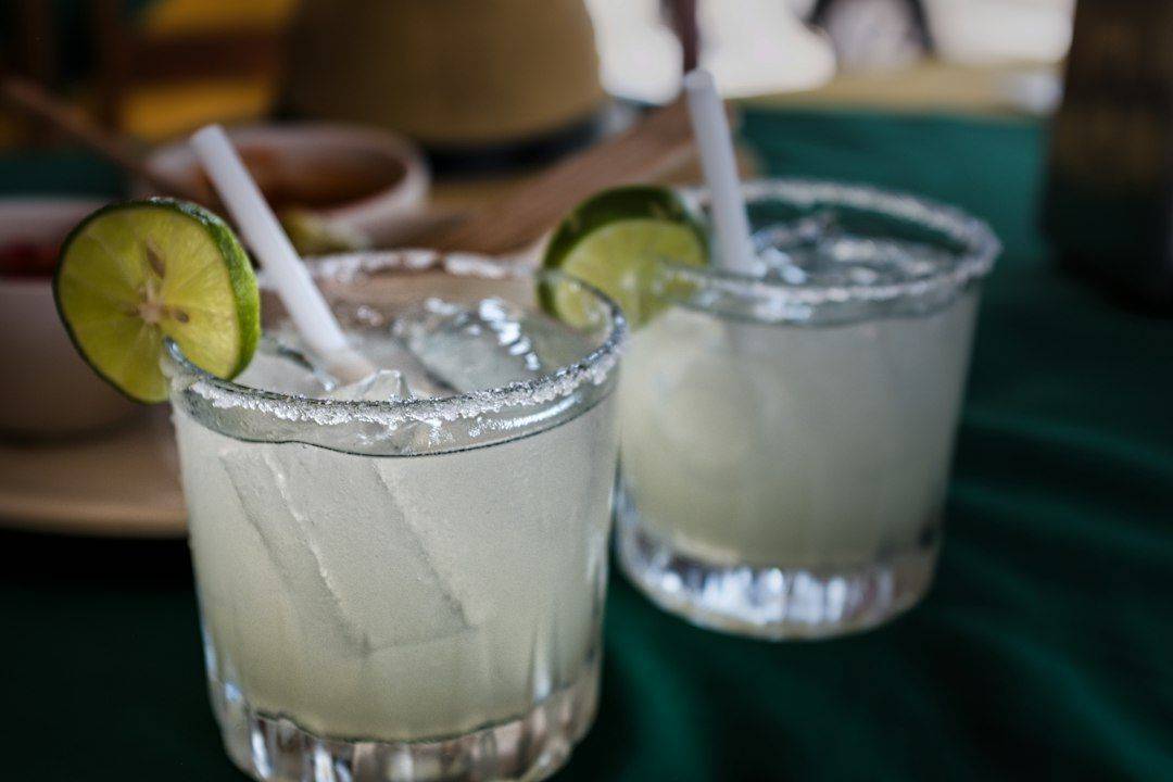 Two Salted-Rim Margaritas With Lime Wedges on Green Tablecloth - A close-up shot of two clear glasses filled with refreshing margaritas. They have salted rims and are garnished with lime wedges. One has a straw in it. The glasses are sitting on a green tablecloth, and there are blurry details of other items in the background. (photographie d’illustration)