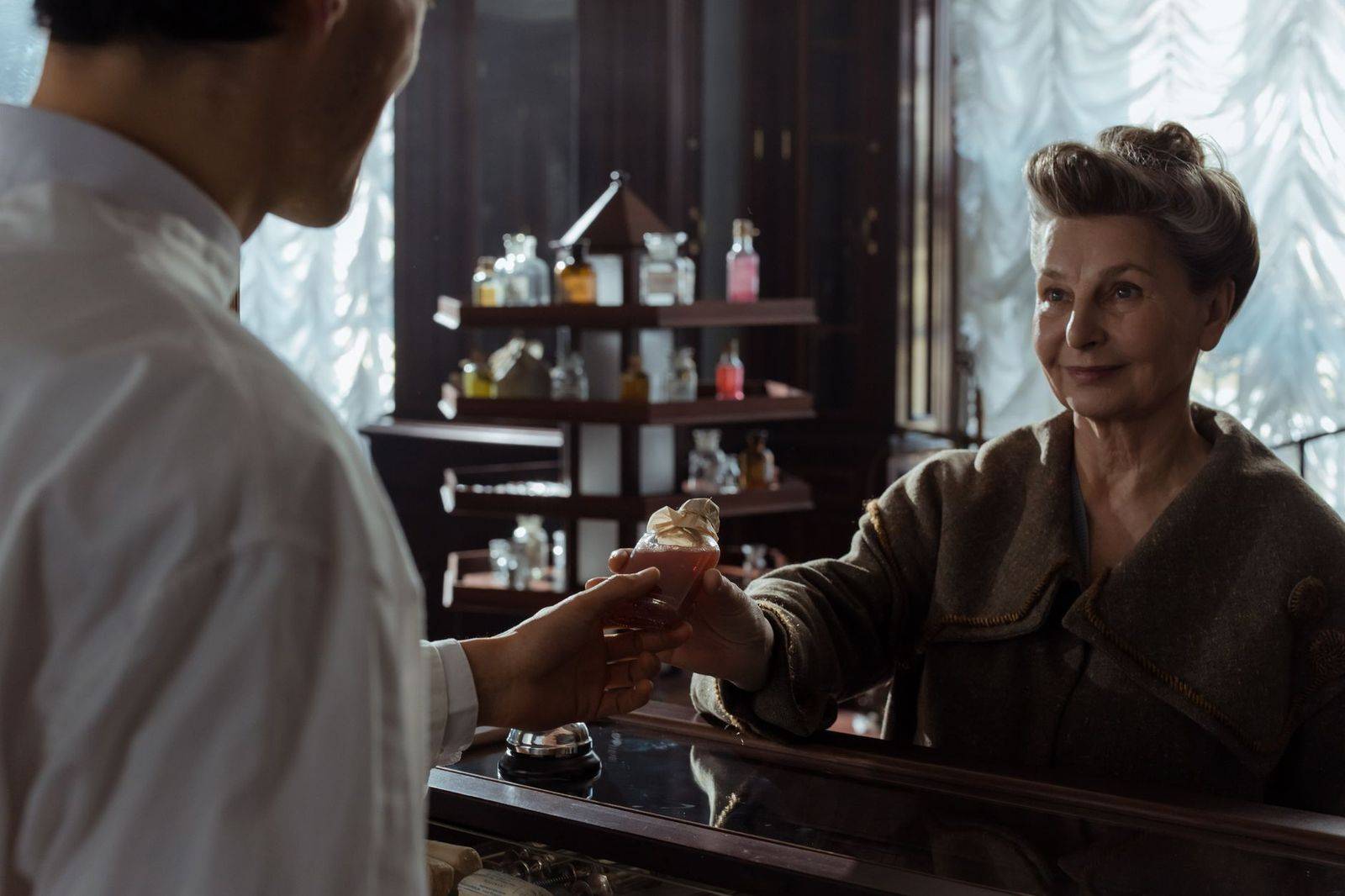 A senior woman receives a perfume bottle from a pharmacist inside a vintage pharmacy. (photographie d’illustration)